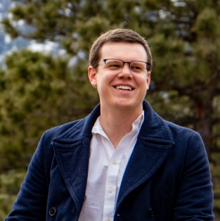 Man in blue coat smiling outdoors with trees in background